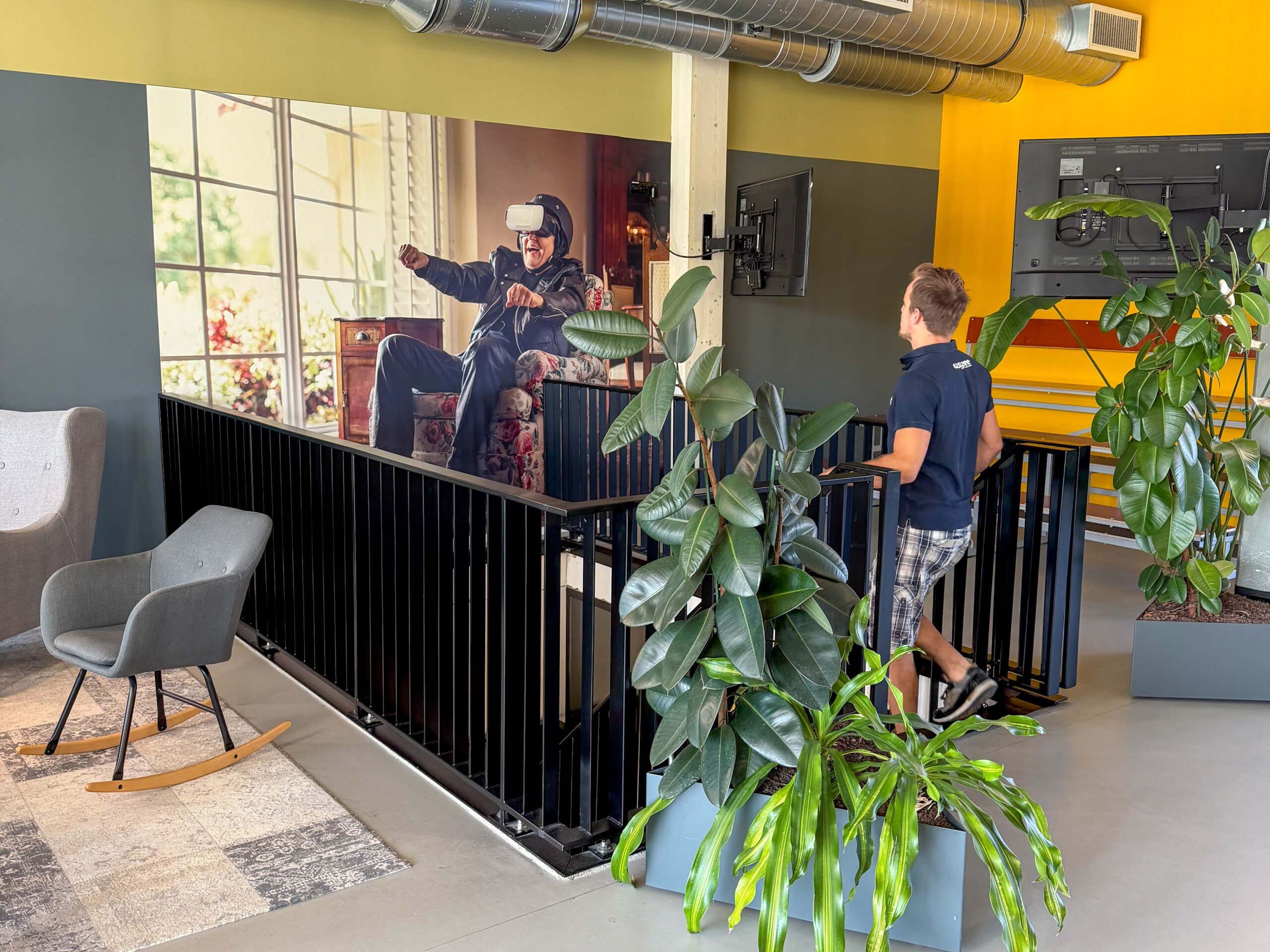 A team member walks up the office stairs past large plants and a VR wall graphic, preparing layout plans for the new VR Experience Room.